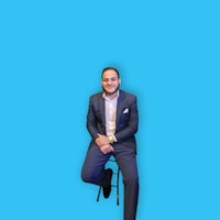 a man in a suit sitting on a stool against a blue background